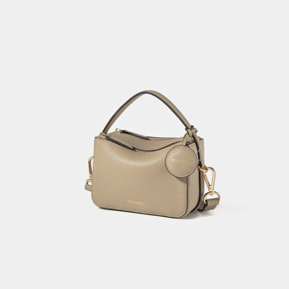 A side-front view of the Mini Double Zip Bag in beige color, made from pebbled leather. The bag features a top handle, gold-tone hardware, and an adjustable shoulder strap. It has two zippered compartments and a small round leather tag attached to the side, embossed with the 'MOONSHAFT' logo in gold. The clean and structured design showcases its minimalist elegance and functional style.