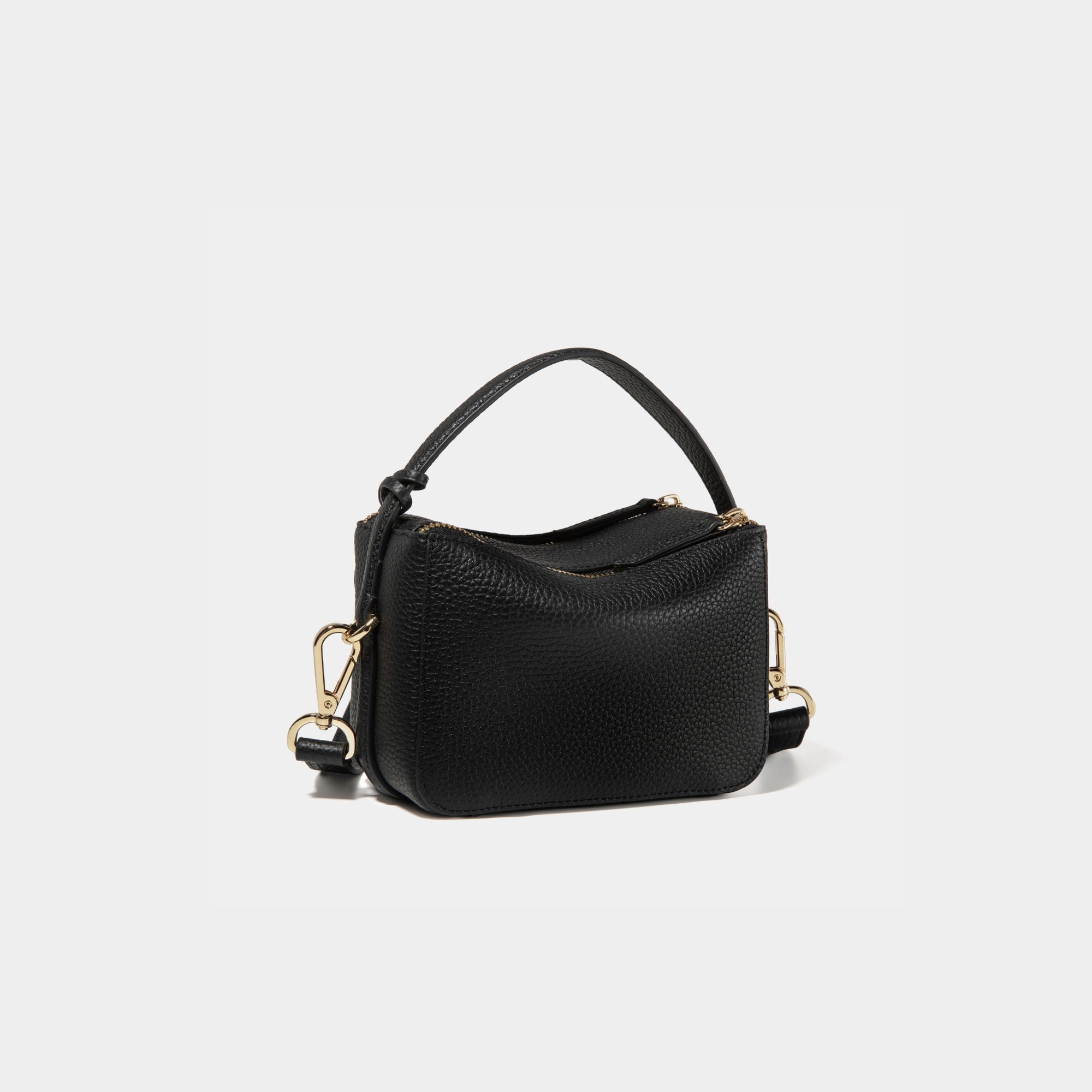 A diagonal back view of the Mini Double Zip Bag in black color, crafted from pebbled leather. The bag features a smooth and structured design with a top handle and detachable gold-tone hardware. The clean back panel showcases the minimalist style, while the adjustable shoulder strap and two zippered compartments add both functionality and elegance to the overall look.