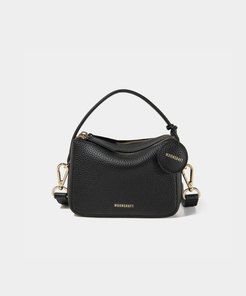 A close-up of the Mini Double Zip Bag in black color, made from pebbled leather. The bag features a top handle and an adjustable shoulder strap with gold-tone hardware. It has two zippered compartments for storage and a round leather tag attached to the side, embossed with the brand logo 'MOONSHAFT' in gold. The minimalist and structured design enhances its modern and stylish appeal.