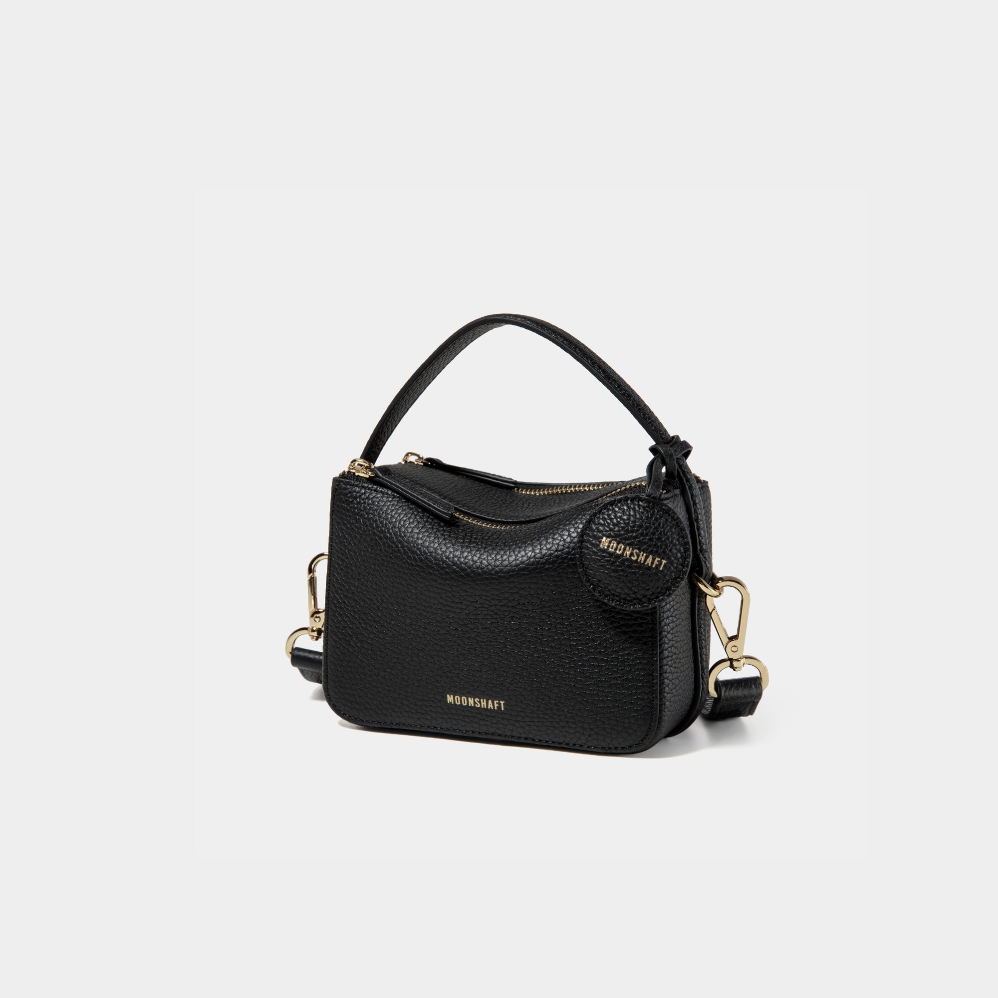 A side-front view of the Mini Double Zip Bag in black color, made from pebbled leather. The bag features a top handle, gold-tone hardware, and an adjustable shoulder strap. It has two zippered compartments and a small round leather tag attached to the side, embossed with the 'MOONSHAFT' logo in gold. The clean and structured design showcases its minimalist elegance and functional style.