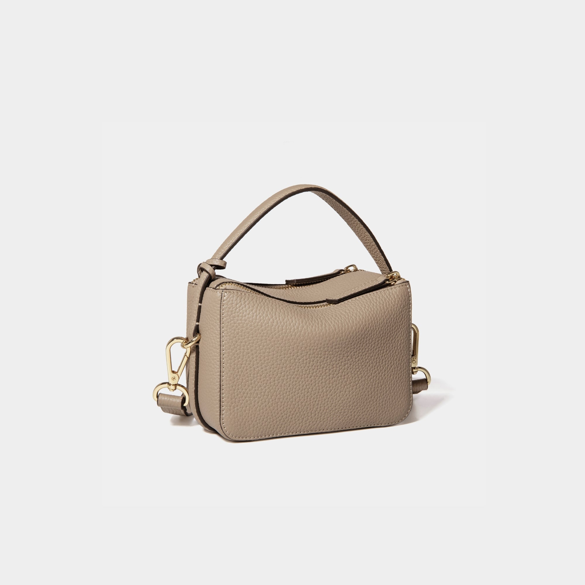 A diagonal back view of the Mini Double Zip Bag in sandstone color, crafted from pebbled leather. The bag features a smooth and structured design with a top handle and detachable gold-tone hardware. The clean back panel showcases the minimalist style, while the adjustable shoulder strap and two zippered compartments add both functionality and elegance to the overall look.