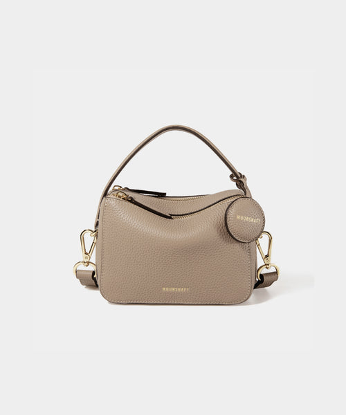 A close-up of the Mini Double Zip Bag in a sandstone color, made from pebbled leather. The bag features a top handle and an adjustable shoulder strap with gold-tone hardware. It has two zippered compartments for storage and a round leather tag attached to the side, embossed with the brand logo 'MOONSHAFT' in gold. The minimalist and structured design enhances its modern and stylish appeal.