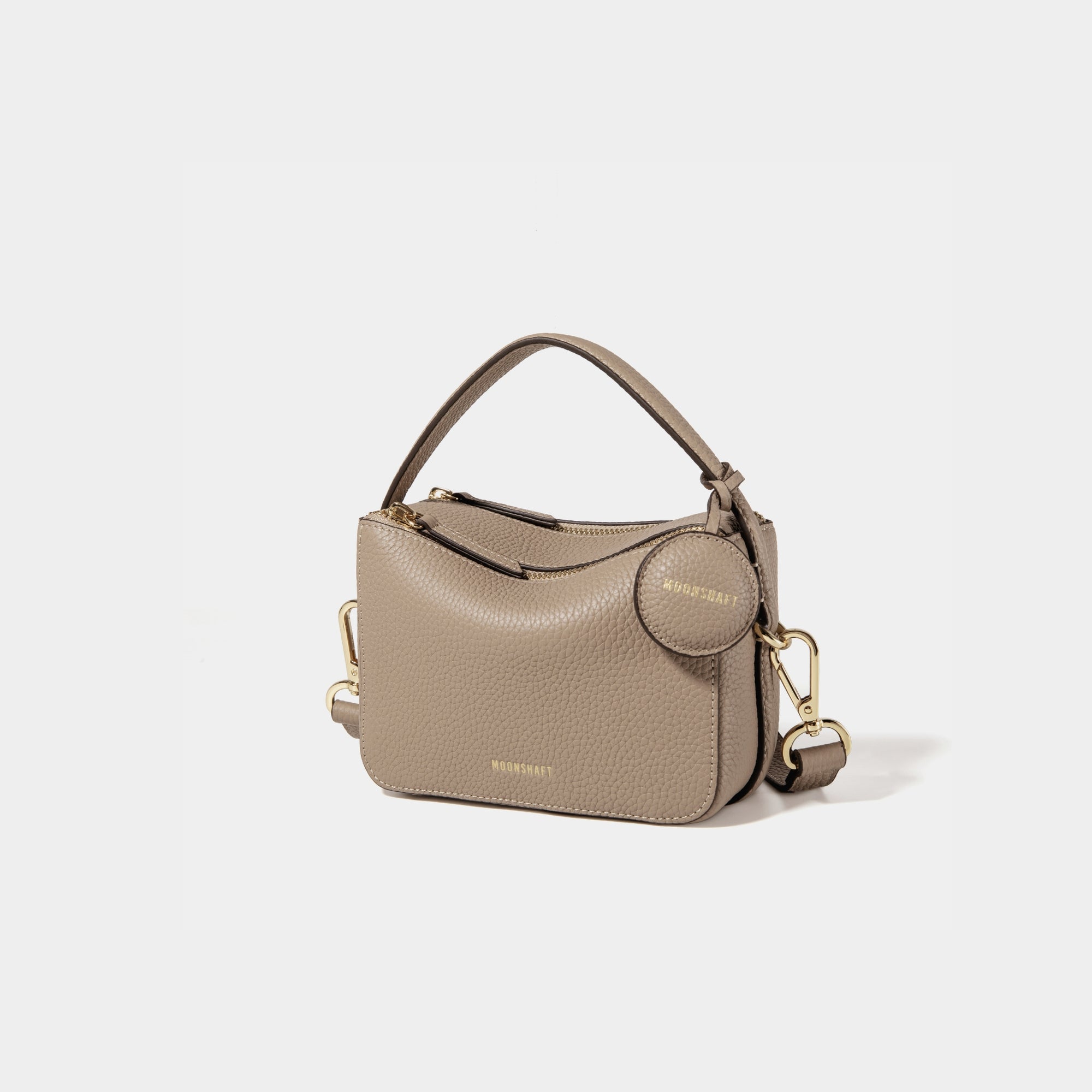 A side-front view of the Mini Double Zip Bag in sandstone color, made from pebbled leather. The bag features a top handle, gold-tone hardware, and an adjustable shoulder strap. It has two zippered compartments and a small round leather tag attached to the side, embossed with the 'MOONSHAFT' logo in gold. The clean and structured design showcases its minimalist elegance and functional style.