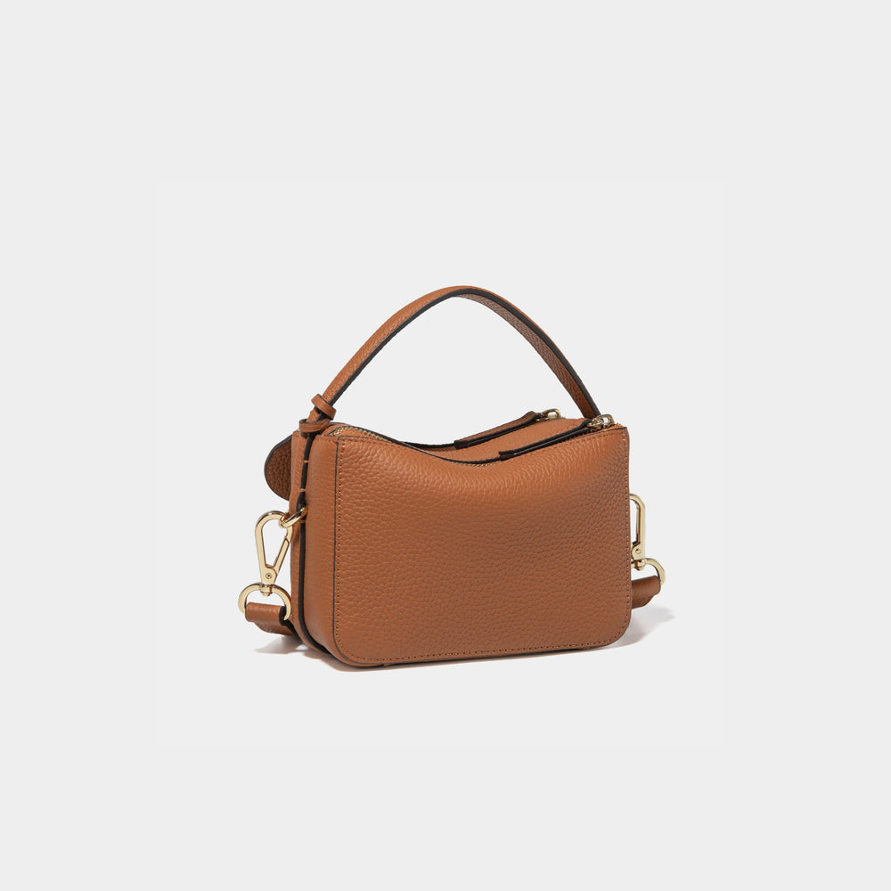 A diagonal back view of the Mini Double Zip Bag in toffee color, crafted from pebbled leather. The bag features a smooth and structured design with a top handle and detachable gold-tone hardware. The clean back panel showcases the minimalist style, while the adjustable shoulder strap and two zippered compartments add both functionality and elegance to the overall look.