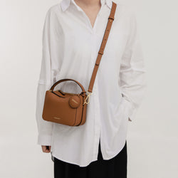 A model wearing a white oversized shirt and black pants carries the Mini Double Zip Bag in toffee color as a crossbody. The bag, made from pebbled leather, features a gold-tone adjustable strap, a top handle, and a round leather tag embossed with the 'MOONSHAFT' logo. The structured and minimalist design complements the model's casual yet stylish outfit.