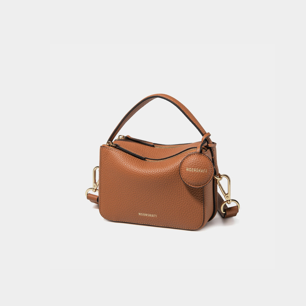 A side-front view of the Mini Double Zip Bag in toffee color, made from pebbled leather. The bag features a top handle, gold-tone hardware, and an adjustable shoulder strap. It has two zippered compartments and a small round leather tag attached to the side, embossed with the 'MOONSHAFT' logo in gold. The clean and structured design showcases its minimalist elegance and functional style.