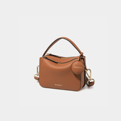 A side-front view of the Mini Double Zip Bag in toffee color, made from pebbled leather. The bag features a top handle, gold-tone hardware, and an adjustable shoulder strap. It has two zippered compartments and a small round leather tag attached to the side, embossed with the 'MOONSHAFT' logo in gold. The clean and structured design showcases its minimalist elegance and functional style.