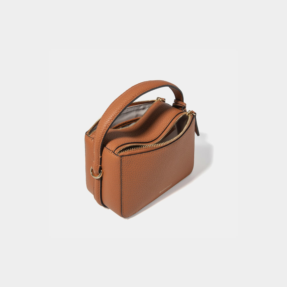 A top diagonal back view of the Mini Double Zip Bag in toffee color, made from pebbled leather. The smooth, structured back panel is visible, highlighting the minimalist design. The bag features two zippered compartments with a light-colored lining, a sturdy top handle, and gold-tone hardware, combining functionality with a sleek, elegant appearance.