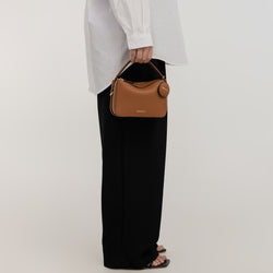 A model dressed in a loose white shirt and black pants holds the Mini Double Zip Bag in toffee color by its top handle. The bag, made from pebbled leather, features a small round leather tag and gold-tone hardware. The minimalist and structured design of the bag complements the model’s casual outfit, showcasing its versatility and chic style.