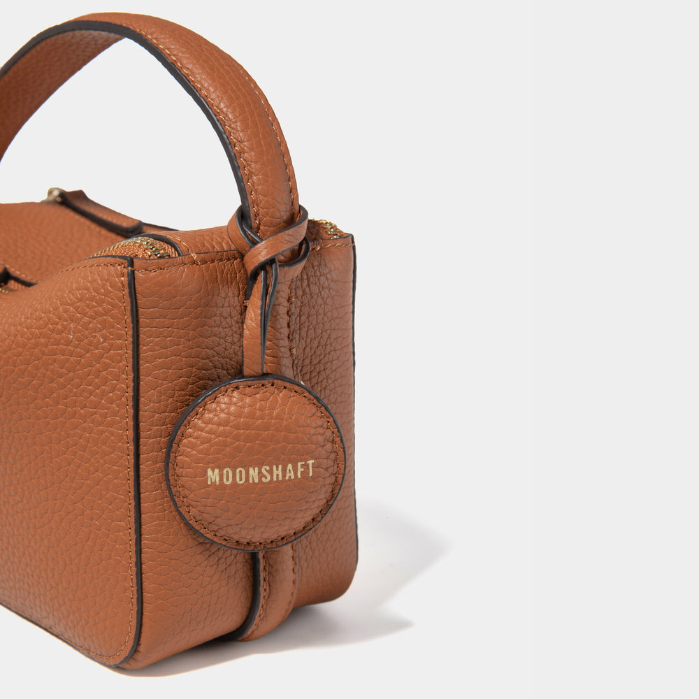 A close-up side view of the Mini Double Zip Bag in toffee color, featuring a detailed look at the round leather tag embossed with the 'MOONSHAFT' logo in gold. The pebbled leather texture and stitching on the bag and handle are highlighted, showcasing the craftsmanship. The structured design and attention to detail enhance the luxurious aesthetic of the bag.
