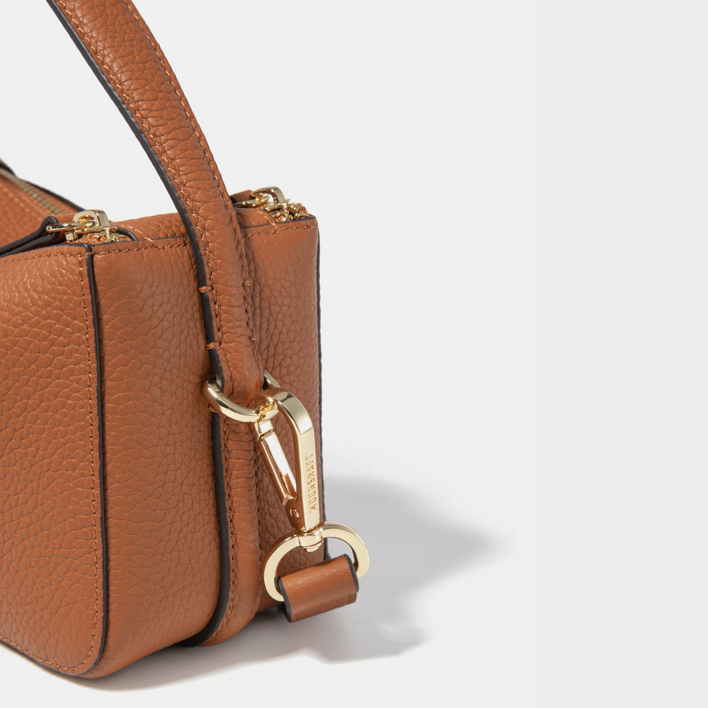 A close-up detail of the Mini Double Zip Bag in toffee color, highlighting the pebbled leather texture and gold-tone hardware. The focus is on the bag's side, showing the detachable strap with a gold-tone clip, engraved with the 'MOONSHAFT' logo. The stitching and high-quality craftsmanship are clearly visible, emphasizing the luxury and durability of the design.