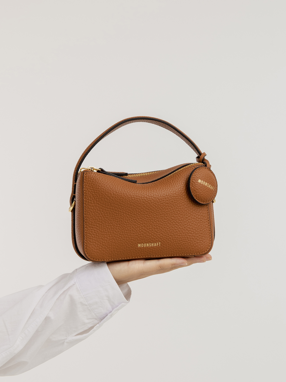 The Mini Double Zip Bag in toffee color is held in a hand, showcasing its compact, structured design. Made from pebbled leather, the bag features a top handle, gold-tone hardware, and a round leather tag embossed with the 'MOONSHAFT' logo. The minimalist and elegant design emphasizes the bag’s versatile and functional style.