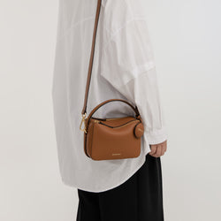 A model wearing a white loose shirt and black trousers is shown carrying a compact crossbody Mini Double Zip bag in toffee color. It has a pebbled texture with a structured design. It features gold-tone hardware, a top handle, a long adjustable strap, and two zippered compartments. A small, round leather tag is attached to the side, adding a stylish accent.