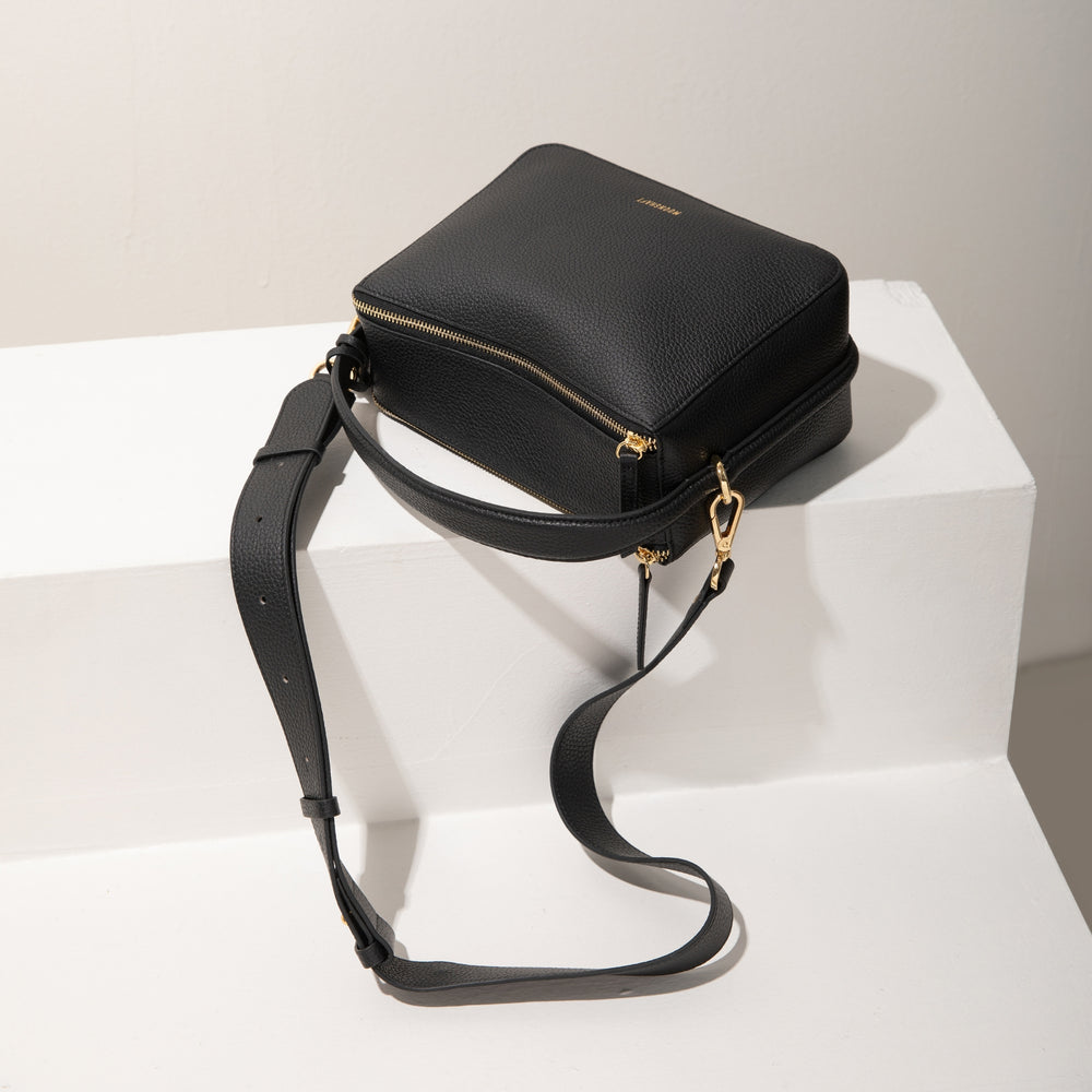 A top view of the MOONSHAFT upgraded double zip pebbled leather bag in black, displayed on a white platform. The bag's structured design is highlighted, along with its gold-tone zippers and hardware. The adjustable, detachable strap is laid out casually, emphasizing the bag’s versatility and functionality. The smooth, pebbled leather texture and clean lines create a chic, minimalist aesthetic, making the bag a stylish accessory for various occasions.