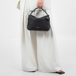 A model holding the MOONSHAFT upgraded double zip pebbled leather bag in black by the top handle. The bag is shown against the backdrop of the model’s white, flowing wide-leg trousers, creating a stylish contrast. The bag's minimalist design is highlighted, featuring gold-tone zippers and a round leather charm with the MOONSHAFT logo. The structured shape and smooth pebbled leather texture make it a chic and versatile accessory for various outfits and occasions