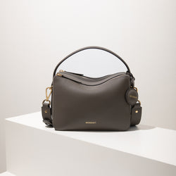 A front view of the MOONSHAFT upgraded double zip pebbled leather bag in charcoal, displayed on a white platform. The bag features a structured design with gold-tone zippers and hardware, including a detachable strap and a top handle for versatility. The pebbled leather texture is highlighted, along with the minimalist MOONSHAFT logo embossed in gold on the front. The round leather charm with the brand’s logo adds a stylish finishing touch, making this bag both elegant and functional.