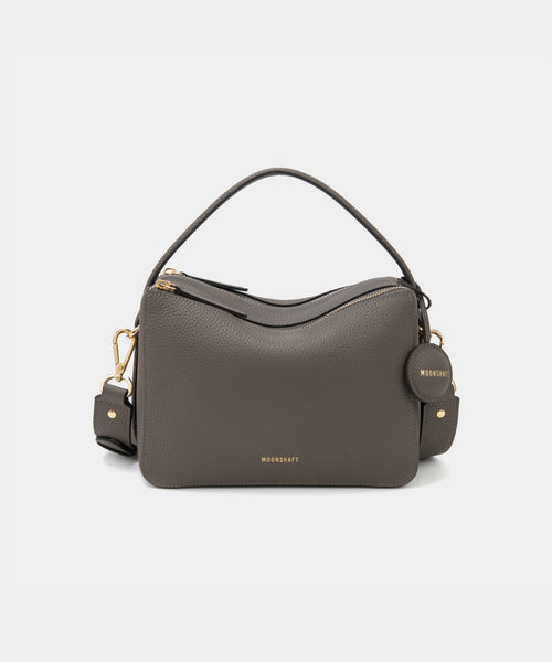 A charcoal-colored upgraded double zip leather bag displayed from a front angle against a plain background. The bag showcases a minimalist design with pebbled leather and two prominent zippered compartments. The structured form and gold-tone zippers add both functionality and a sleek, modern touch. The neutral charcoal color enhances the bag's versatile appeal, making it suitable for a variety of outfits and occasions.