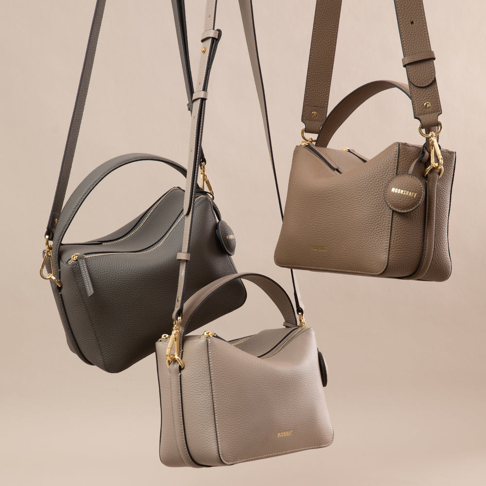 Three MOONSHAFT upgraded double zip pebbled leather bags in neutral tones (charcoal, driftwood, and sandstone) are stylishly suspended by their straps. Each bag showcases a structured design with gold-tone zippers and hardware, along with a top handle and an adjustable shoulder strap. The smooth pebbled leather texture and minimalist aesthetic highlight the elegance and versatility of the bags, making them perfect for various outfits and occasions. The neutral background complements the sophisticated color 