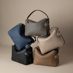 A display of five upgraded double zip pebbled leather bags, artfully stacked in a pyramid-like arrangement. The black bag is positioned at the bottom left, with the marine blue bag leaning diagonally on top of it. To the right, the driftwood bag sits at the bottom, while the sandstone bag rests above it, slightly tilted forward. At the top, the charcoal bag is balanced, showcasing its structured form. The pebbled leather texture and gold-tone zippers are highlighted, offering a chic and versatile look.