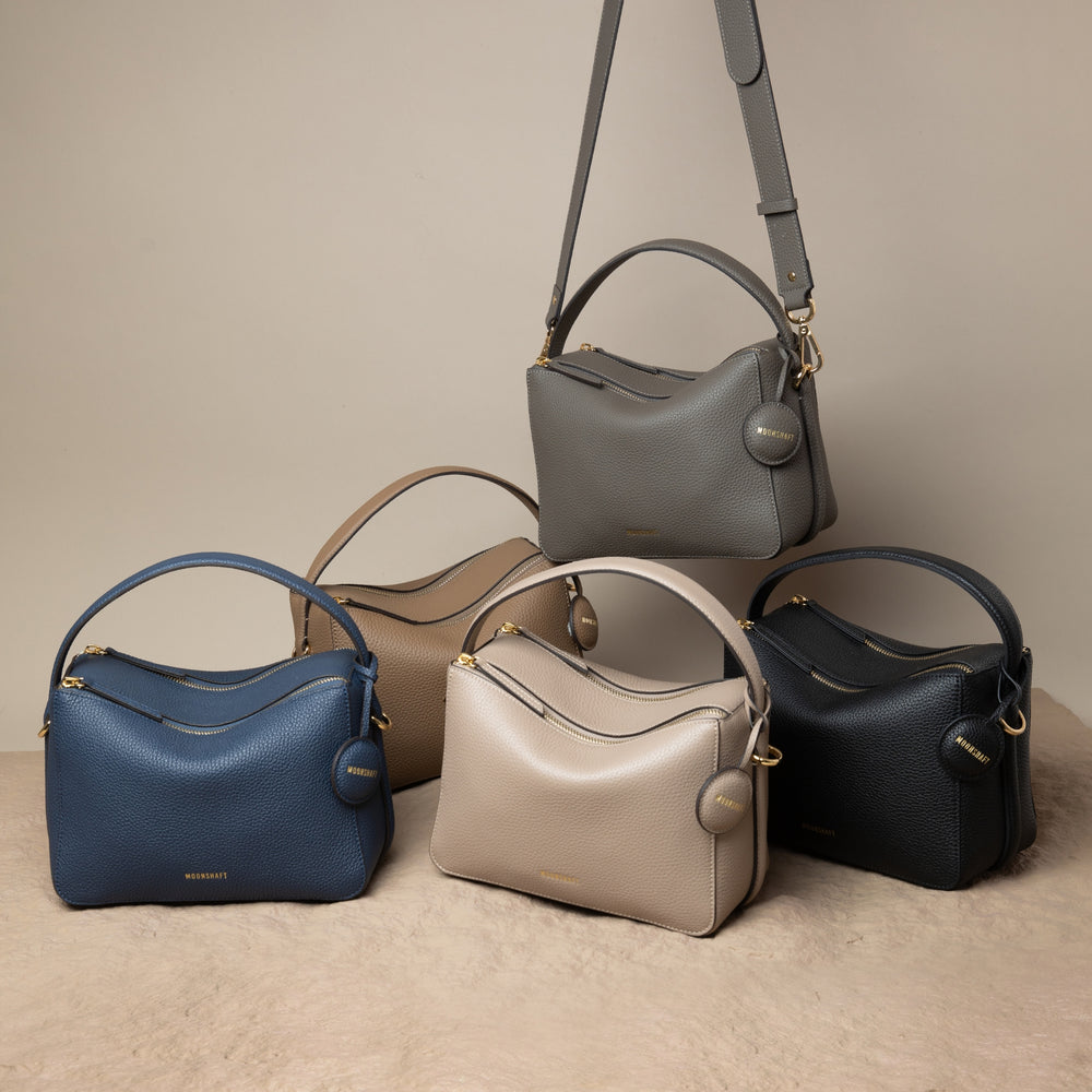 A display of the upgrade double zip bags in various colors: marine blue, driftwood, sandstone, charcoal, and black. One of the charcoal bags is shown hanging by its adjustable strap, while the other four are arranged neatly on a beige textured surface. Each bag features a structured, minimalist design with pebbled leather, gold-tone zippers, and a single top handle. The MOONSHAFT logo is embossed on the front, and a round leather charm is attached to each bag, showcasing their elegant yet functional 