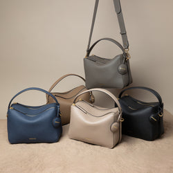 A display of the upgrade double zip bags in various colors: marine blue, driftwood, sandstone, charcoal, and black. One of the charcoal bags is shown hanging by its adjustable strap, while the other four are arranged neatly on a beige textured surface. Each bag features a structured, minimalist design with pebbled leather, gold-tone zippers, and a single top handle. The MOONSHAFT logo is embossed on the front, and a round leather charm is attached to each bag, showcasing their elegant yet functional 