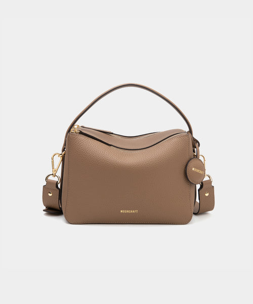 A driftwood-colored upgraded double zip leather bag displayed from a front angle against a plain background. The bag showcases a minimalist design with pebbled leather and two prominent zippered compartments. The structured form and gold-tone zippers add both functionality and a sleek, modern touch. The neutral driftwood color enhances the bag's versatile appeal, making it suitable for a variety of outfits and occasions.