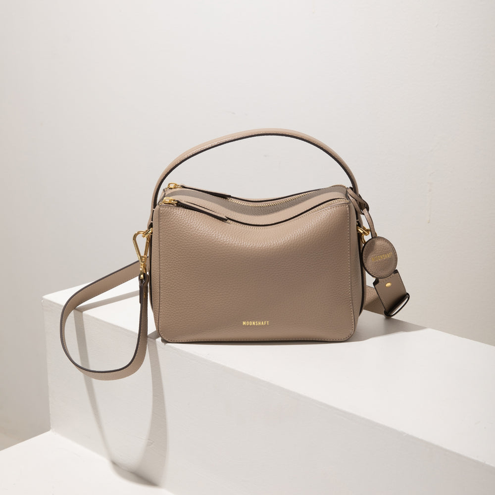 A front view of the upgraded double zip leather bag in sandstone, elegantly displayed on a white platform. The bag features a structured design with smooth, pebbled leather, gold-tone zippers, and hardware. The MOONSHAFT logo is embossed in gold on the front, and a circular leather charm with the logo hangs from the side. The detachable strap is casually draped to the side, emphasizing the bag's functionality. The soft sandstone color adds a refined, minimalist aesthetic, perfect for various occasions.