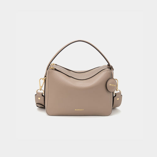 A sandstone-colored upgraded double zip leather bag displayed from a front angle against a plain background. The bag showcases a minimalist design with pebbled leather and two prominent zippered compartments. The structured form and gold-tone zippers add both functionality and a sleek, modern touch. The neutral sandstone color enhances the bag's versatile appeal, making it suitable for a variety of outfits and occasions.