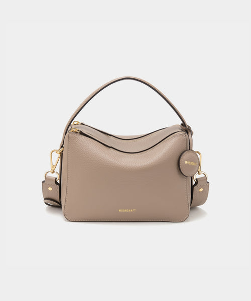 A sandstone-colored upgraded double zip leather bag displayed from a front angle against a plain background. The bag showcases a minimalist design with pebbled leather and two prominent zippered compartments. The structured form and gold-tone zippers add both functionality and a sleek, modern touch. The neutral sandstone color enhances the bag's versatile appeal, making it suitable for a variety of outfits and occasions.