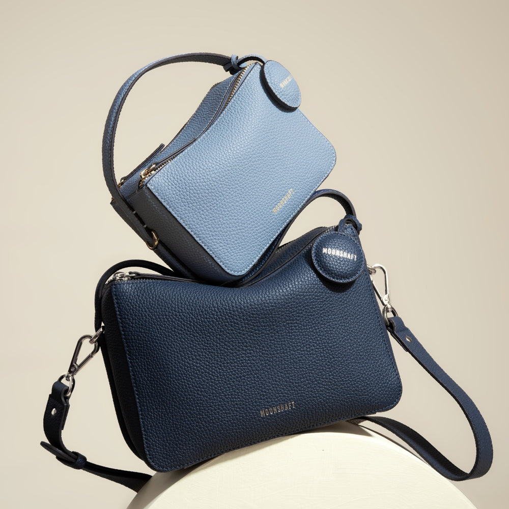 A display of two MOONSHAFT bags, the Mini Double Zip Bag in aquamarine, and the larger Original Double Zip Bag in marine blue. Both bags are made from pebbled leather and feature gold-tone hardware, two zippered compartments, and round leather tags embossed with the 'MOONSHAFT' logo. The structured and minimalist design of both bags emphasizes their modern elegance and functionality.