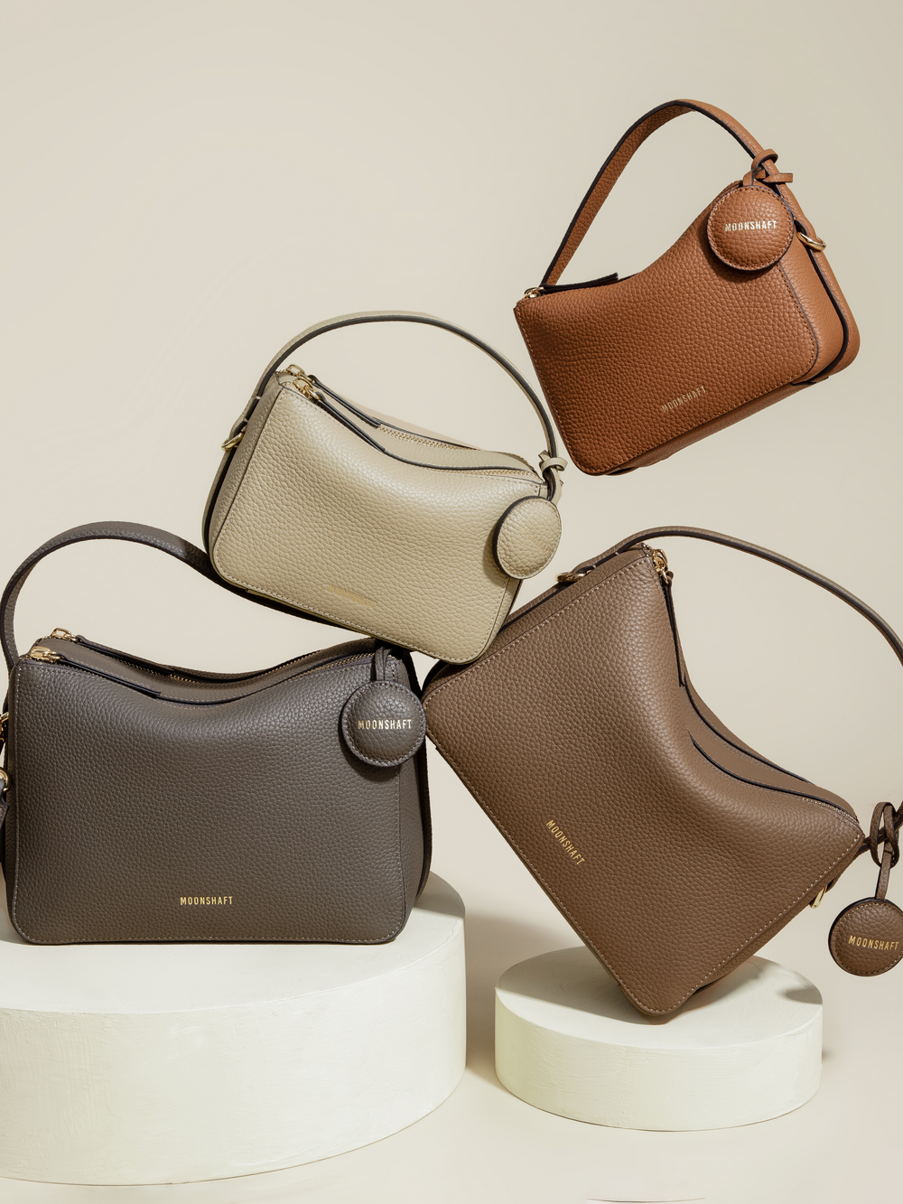 A creative display of four MOONSHAFT bags in various neutral tones, including toffee, beige, taupe, and brown. The arrangement shows both Mini Double Zip Bags and Original Double Zip Bags, all made from pebbled leather. Each bag features gold-tone hardware, round leather tags embossed with the 'MOONSHAFT' logo, and structured designs that highlight their elegant, minimalist aesthetic.