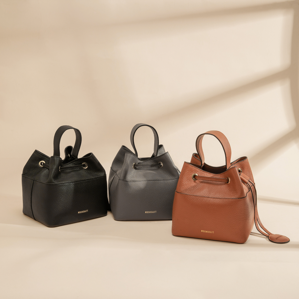 Three premium pebbled leather bucket bags in black, gray, and tan, displayed on a soft beige background. Each bag features a structured silhouette with a top handle, gold-tone hardware, and a drawstring closure for a refined, minimalist look. The bags are elegantly embossed with the MOONSHAFT logo, and the tan bag includes a detachable coin pouch hanging from the drawstring for added versatility.