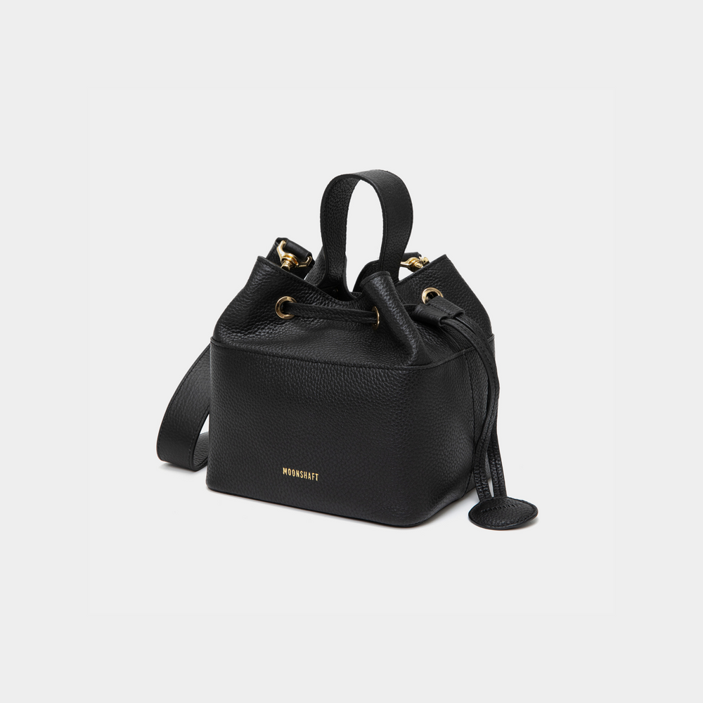 Angled view of a black-colored pebbled leather bucket bag with gold-tone accents, featuring a drawstring closure and a sturdy top handle. The MOONSHAFT logo is embossed in gold on the front, with a small circular leather charm attached to the side for added elegance.