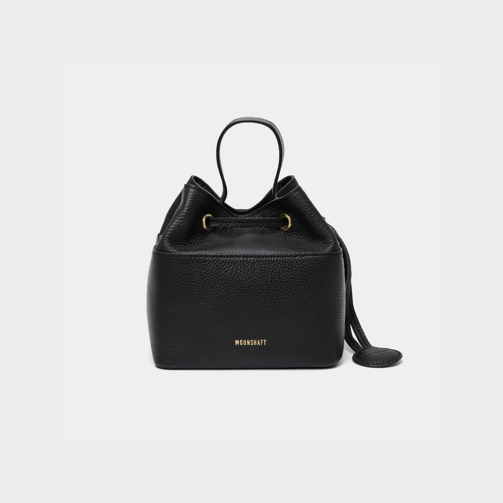 Black-colored pebbled leather bucket bag with gold accents, featuring a drawstring closure and a compact design. The MOONSHAFT logo is embossed in gold at the bottom front. A small round leather accessory is attached to the side for added detail.