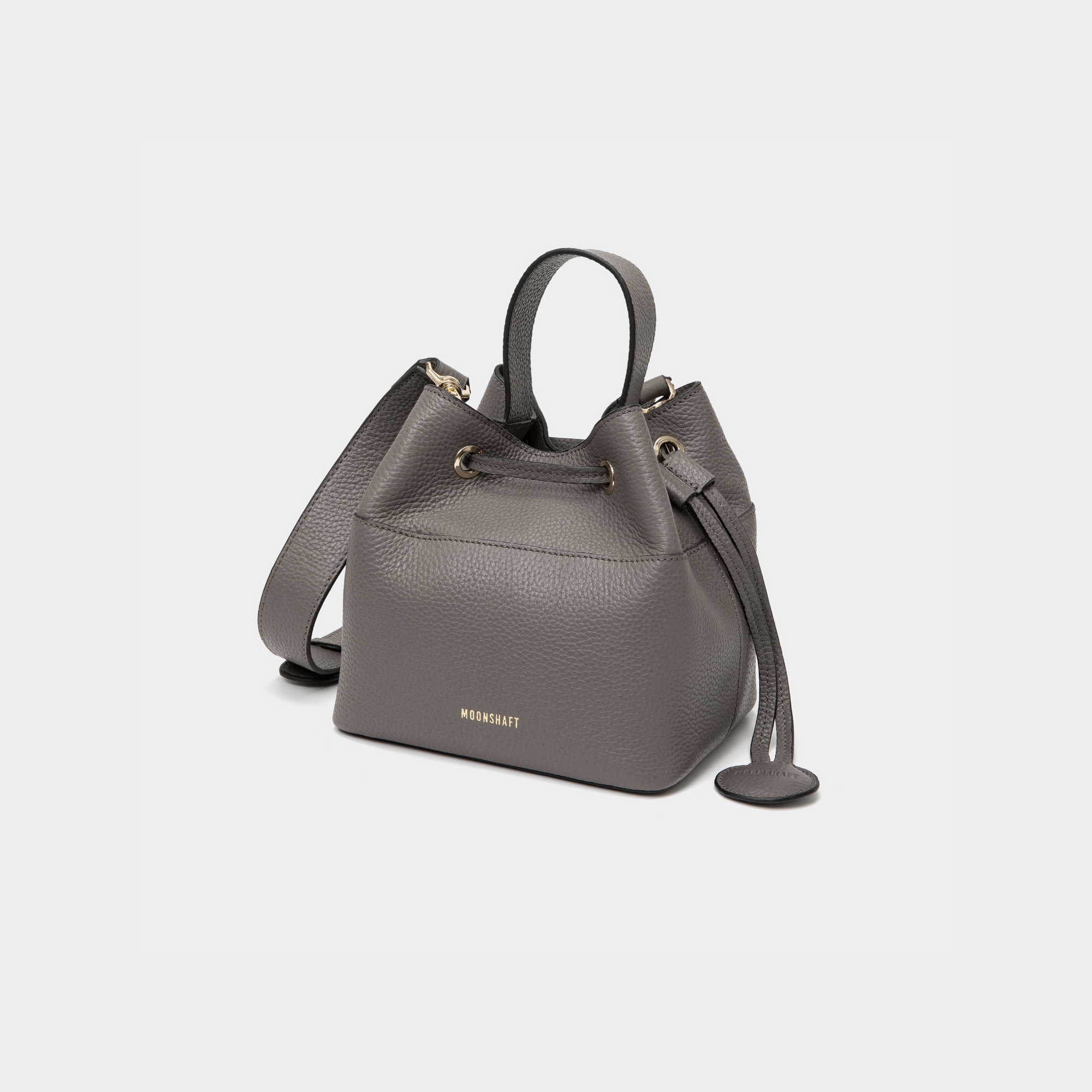 Angled view of a grey-colored pebbled leather bucket bag with gold-tone accents, featuring a drawstring closure and a sturdy top handle. The MOONSHAFT logo is embossed in gold on the front, with a small circular leather charm attached to the side for added elegance.