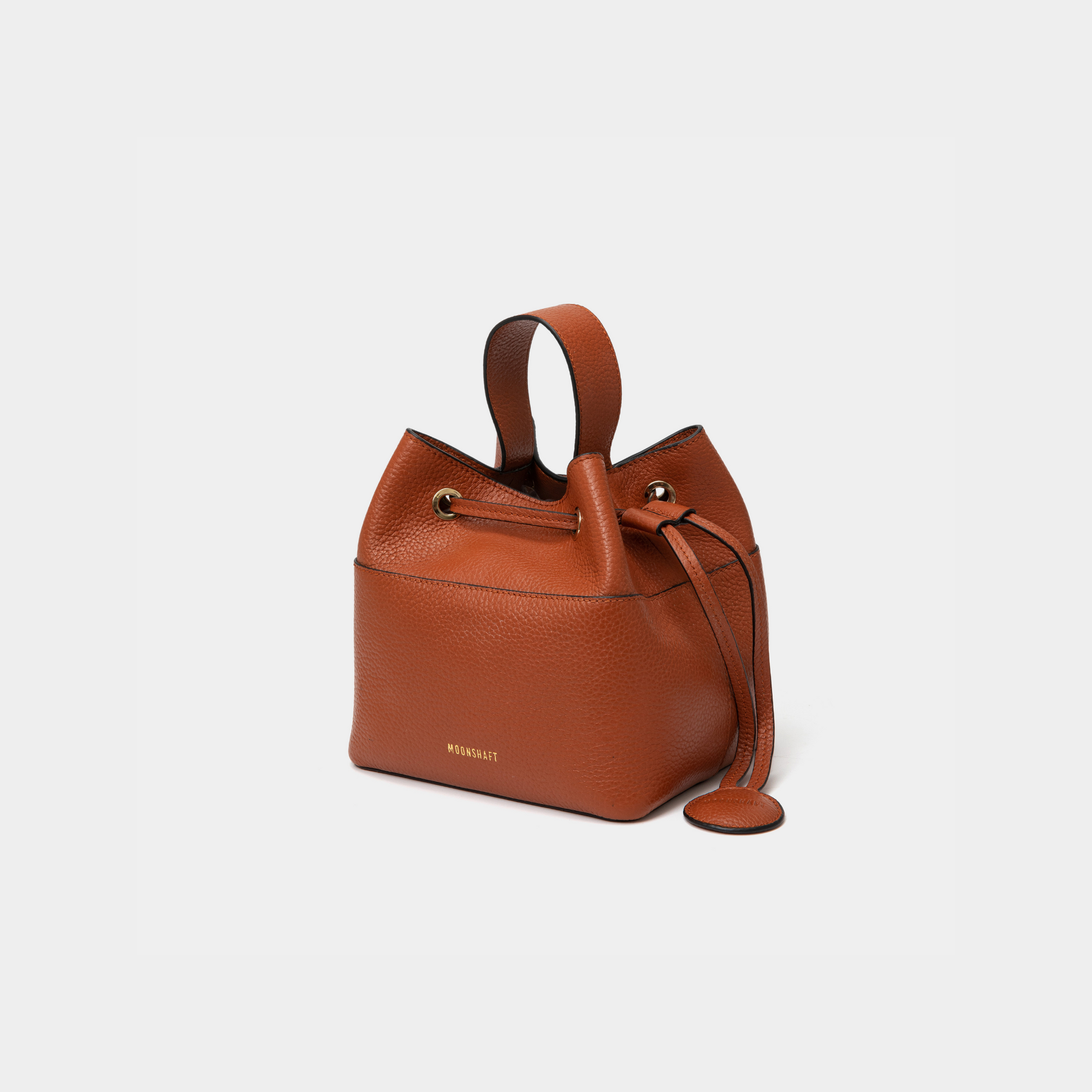 Angled view of a tan-colored pebbled leather bucket bag with gold-tone accents, featuring a drawstring closure and a sturdy top handle. The MOONSHAFT logo is embossed in gold on the front, with a small circular leather charm attached to the side for added elegance.