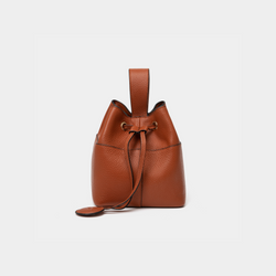 Side view of a tan-colored pebbled leather bucket bag, highlighting the drawstring closure and the sleek top handle. The circular leather charm is attached to the side, adding a stylish touch to the minimalist design.