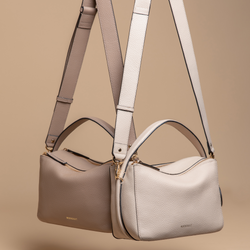 Two MOONSHAFT double zip pebbled leather bags in taupe and sand, suspended by their adjustable straps against a beige background. Each bag features gold-tone zippers, top handles, and detachable straps. The smooth pebbled leather texture and clean design make the bags stand out as elegant and versatile accessories.