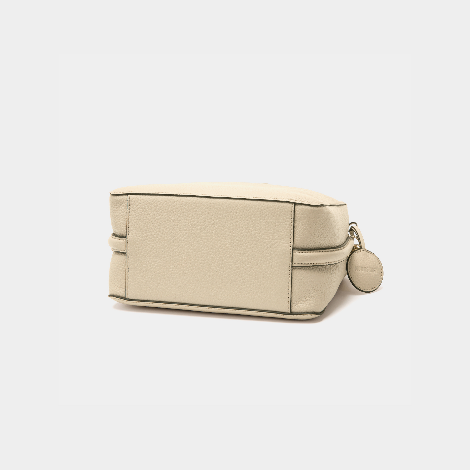 A bottom view of the MOONSHAFT double zip pebbled leather bag in sand, highlighting the structured rectangular base with clean stitching along the edges. The smooth pebbled leather texture is prominent, and the side profile reveals the compact, sleek design of the bag. A glimpse of the attached round leather charm is visible to the side.