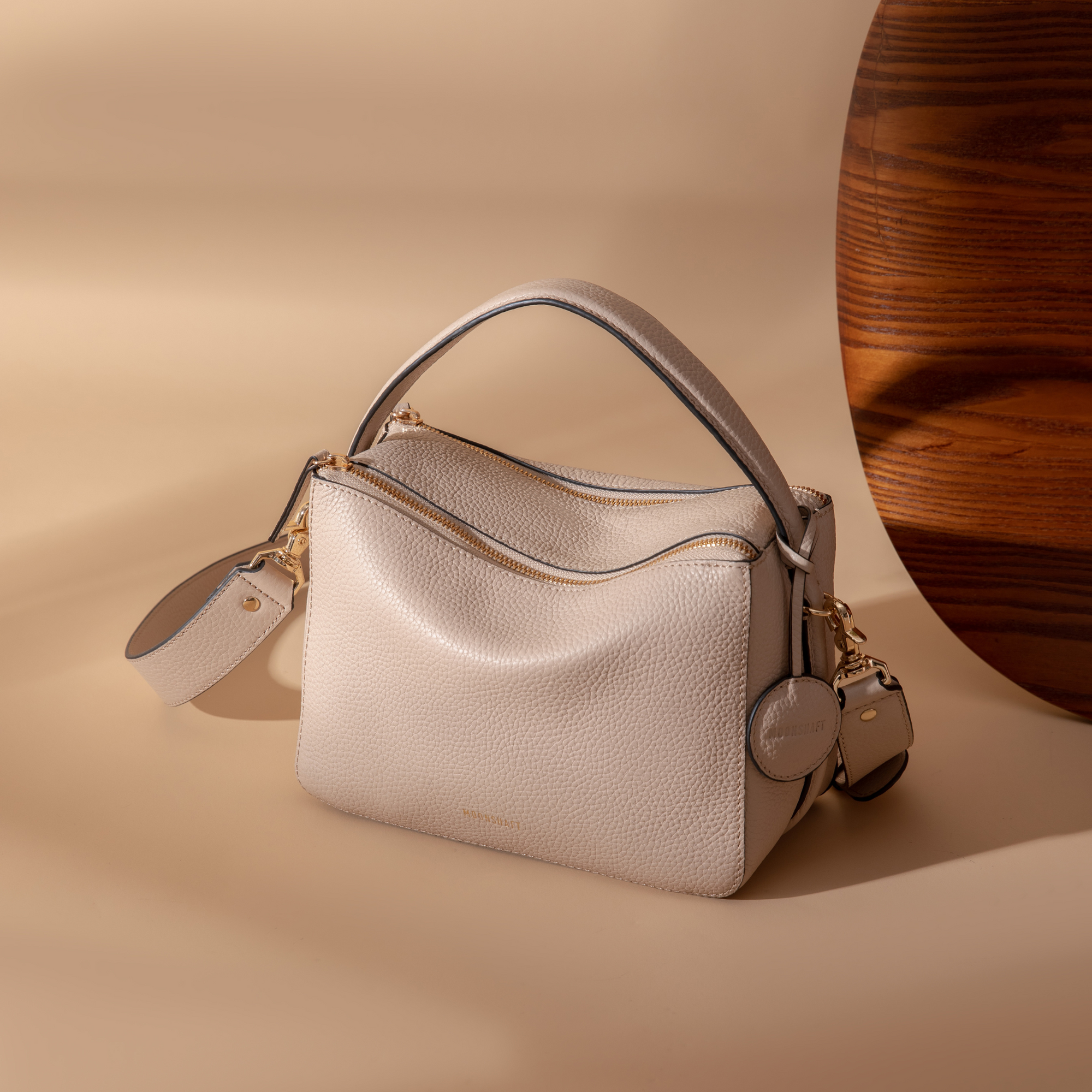 Sand MOONSHAFT double zip pebbled leather bag displayed on a soft beige surface. The bag features gold-tone zippers, a top handle, and a detachable strap. The pebbled leather texture is highlighted, with a round leather charm attached to the side. A wooden accent is visible in the background, adding warmth to the minimalist setup.