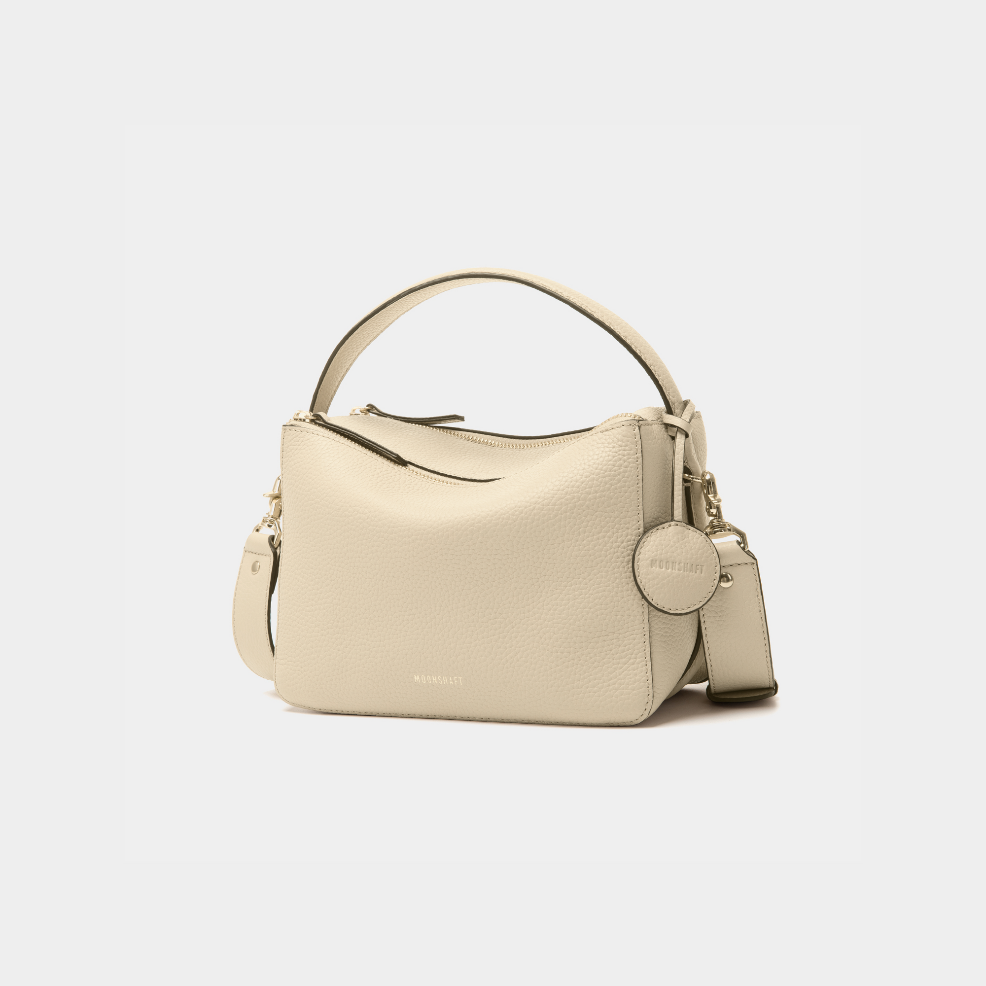 Diagonal view of the MOONSHAFT double zip pebbled leather bag in sand, featuring gold-tone zippers, a top handle, and a detachable strap. The MOONSHAFT logo is embossed in gold on the front, with a round leather charm attached.