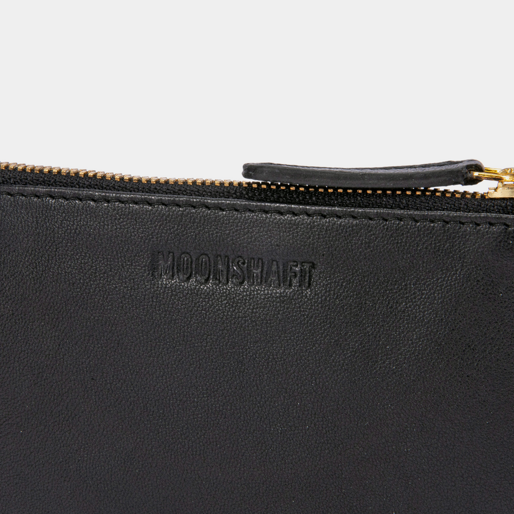 A close-up view of the Half Moon Coin Purse in goat leather, featuring the MOONSHAFT logo embossed on the front. The focus is on the gold-tone zipper and the detailed stitching along the edges of the leather, highlighting the fine craftsmanship. The leather zipper pull is partially visible, accentuating the luxurious texture of the black leather.