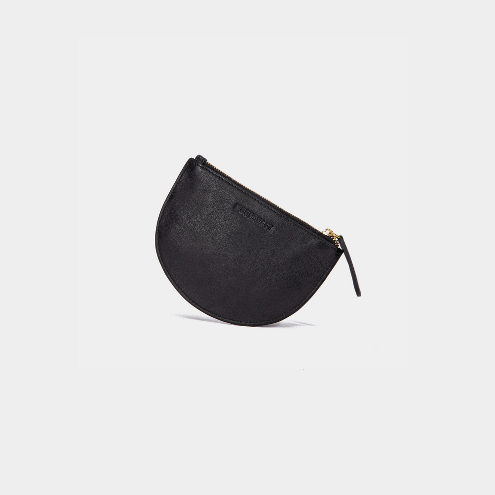 The Half Moon Coin Purse is positioned upright, resting on its curved bottom edge, creating a slight tilt. The zipper is fully closed, and the leather pull tab hangs naturally to the side. The goat leather texture is smooth, with the MOONSHAFT branding subtly embossed near the top center. The product appears balanced, highlighting its slim profile and elegant shape.