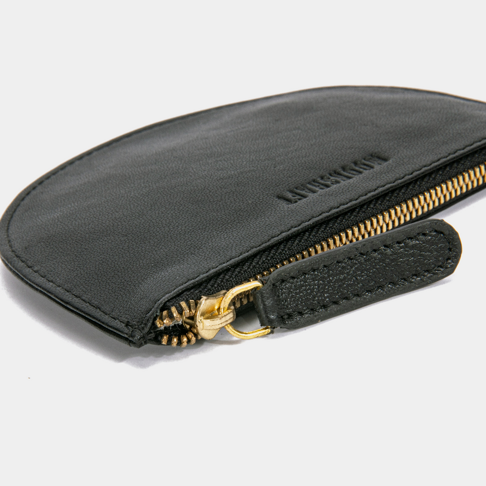 A close-up of the Half Moon Coin Purse, focusing on the gold-tone zipper and leather pull tab. The purse is laid flat with the zipper partially open, showcasing the smooth texture of the goat leather. The MOONSHAFT logo is subtly embossed above the zipper. The stitching along the edges is visible, highlighting the craftsmanship.