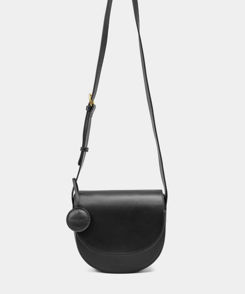 A sleek black half-moon crossbody bag with a curved, structured silhouette. The bag has a smooth leather finish and features a circular leather tag attached to the strap. The adjustable leather strap with a gold-tone buckle adds a touch of sophistication to the minimalist design.