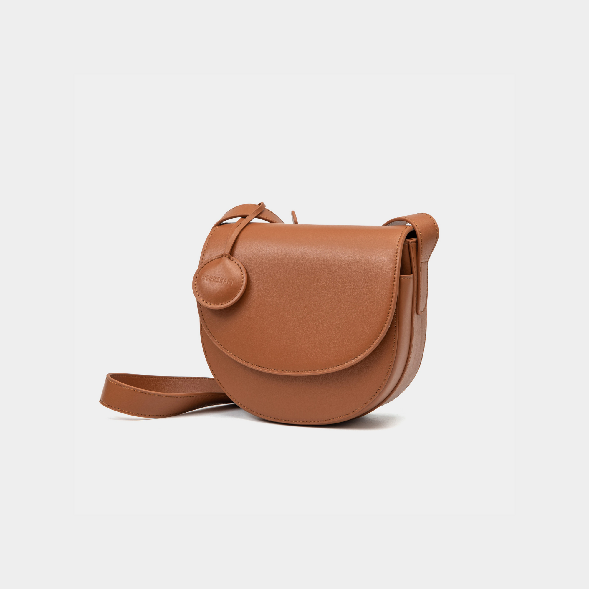A front-side angle view of a caramel half-moon crossbody bag made of smooth leather. The bag has a curved base and minimalistic flap closure. A circular leather tag is attached to the strap, adding a subtle design detail. The adjustable strap and sleek design offer a versatile, elegant look.