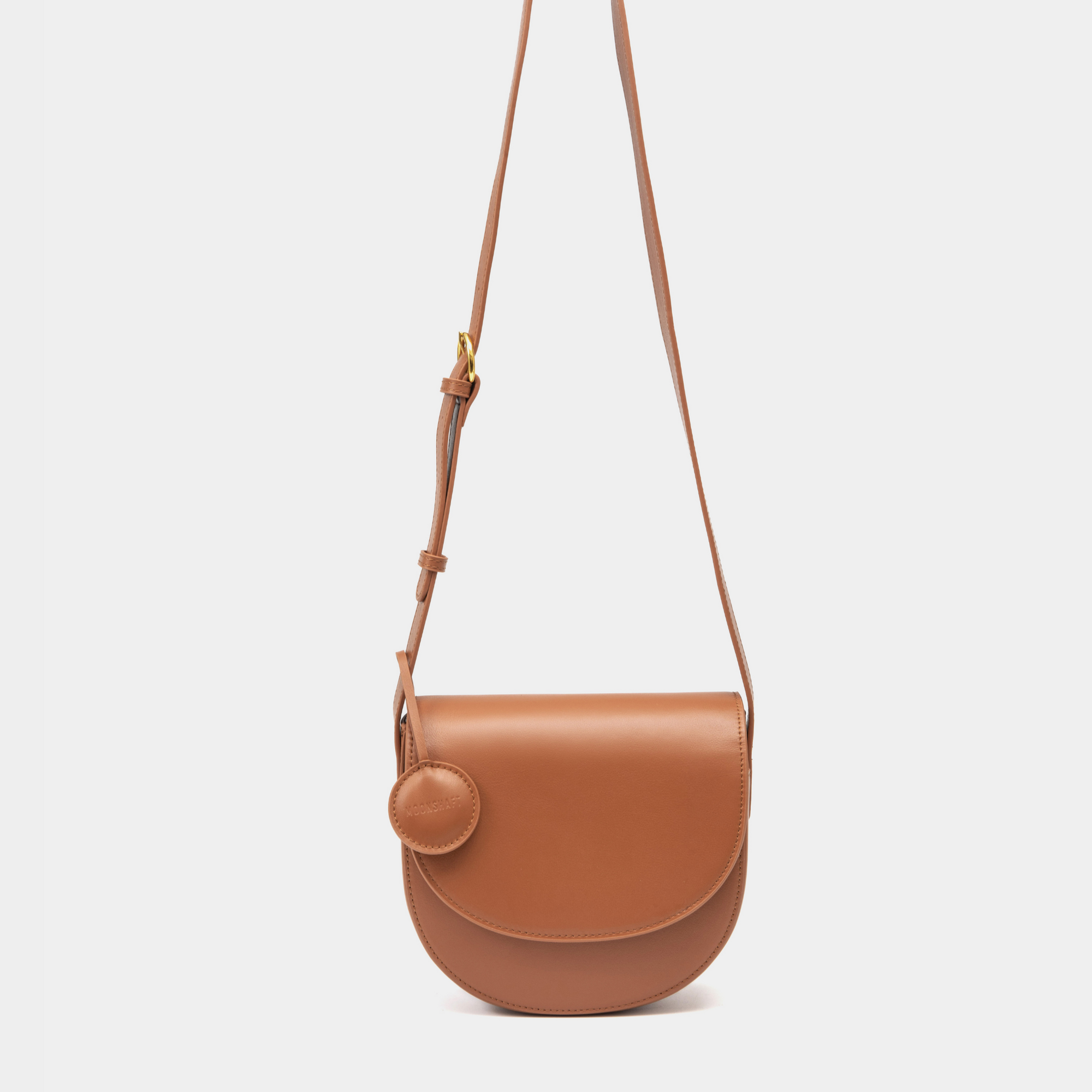 A sleek caramel half-moon crossbody bag with a curved, structured silhouette. The bag has a smooth leather finish and features a circular leather tag attached to the strap. The adjustable leather strap with a gold-tone buckle adds a touch of sophistication to the minimalist design.