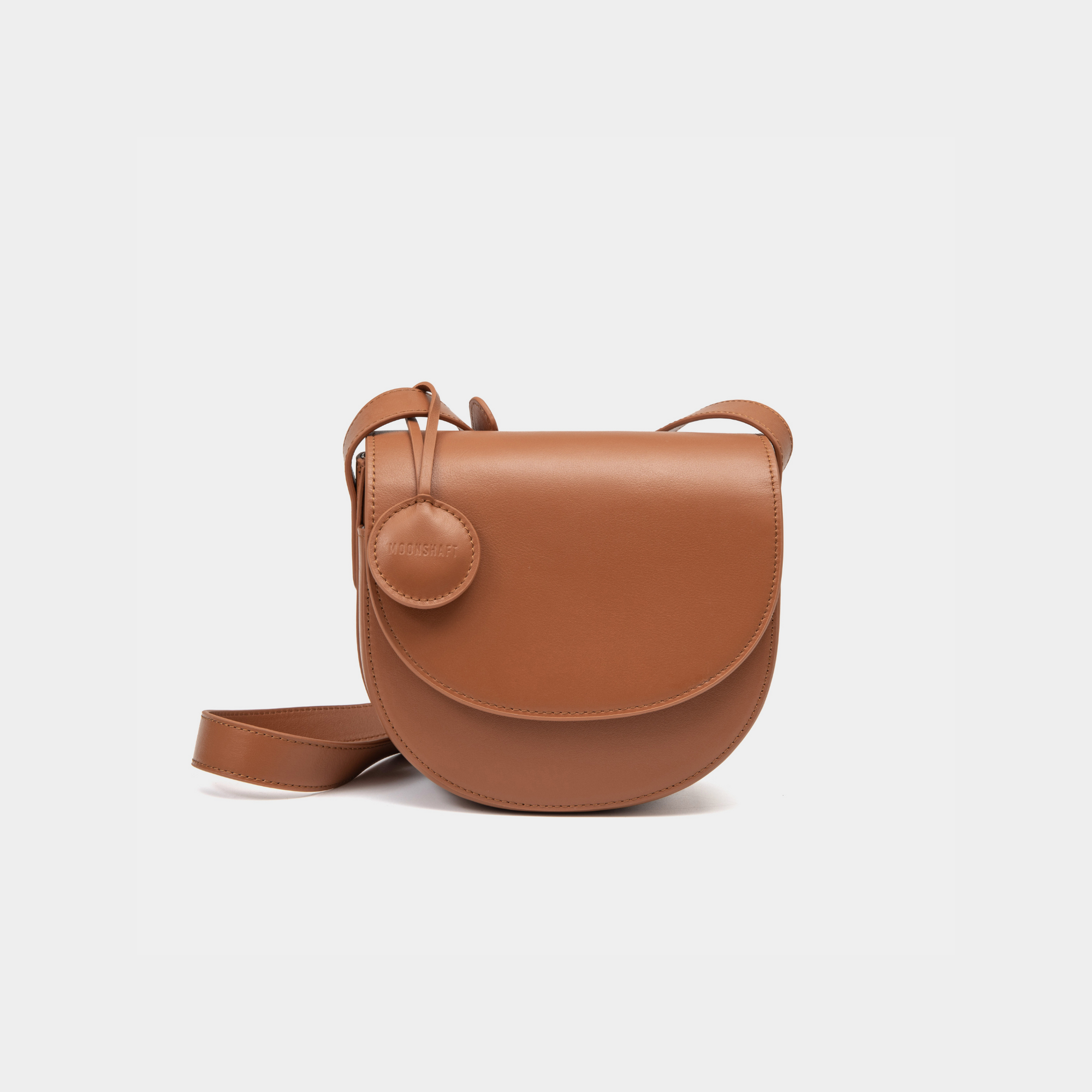 A caramel half-moon crossbody bag with a smooth leather finish, featuring a circular leather tag attached to the strap. The structured design highlights the bag's minimalist aesthetic and sleek curves, offering an elegant yet functional style for everyday wear.