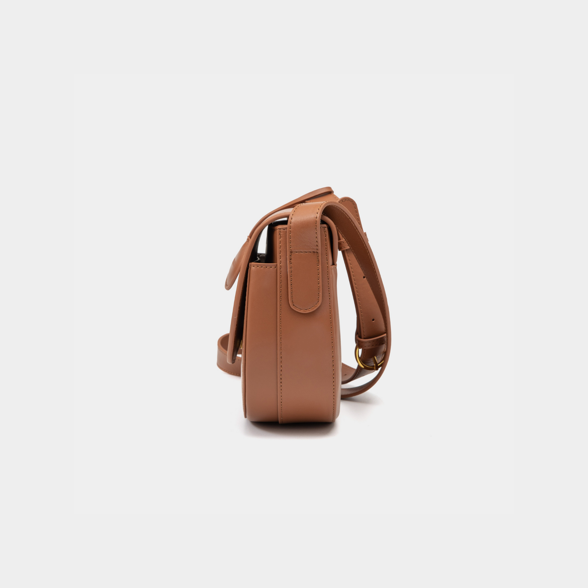 A side view of a caramel half-moon crossbody bag showcasing its structured profile. The bag has a sleek, smooth leather finish, and its curved base complements the minimalistic design. The adjustable leather strap and gold-tone hardware are visible from this angle, adding a touch of refinement.