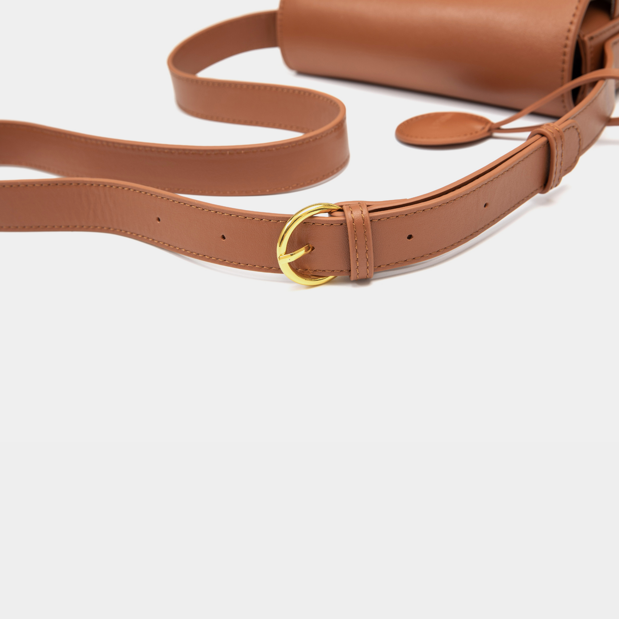 A close-up of a caramel-colored leather crossbody bag strap, showcasing its adjustable buckle design in polished gold. The stitching is neatly aligned along the edges, emphasizing the quality of craftsmanship. The smooth leather strap lies flat, with the circular leather tag in the background, complementing the overall design of the bag.